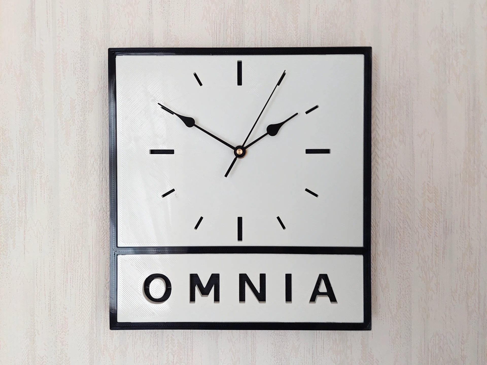 Omnia-logosta inspiroitu 3D-tulostettu seinäkello, musta kehys, valkoinen taulu, musta tausta.