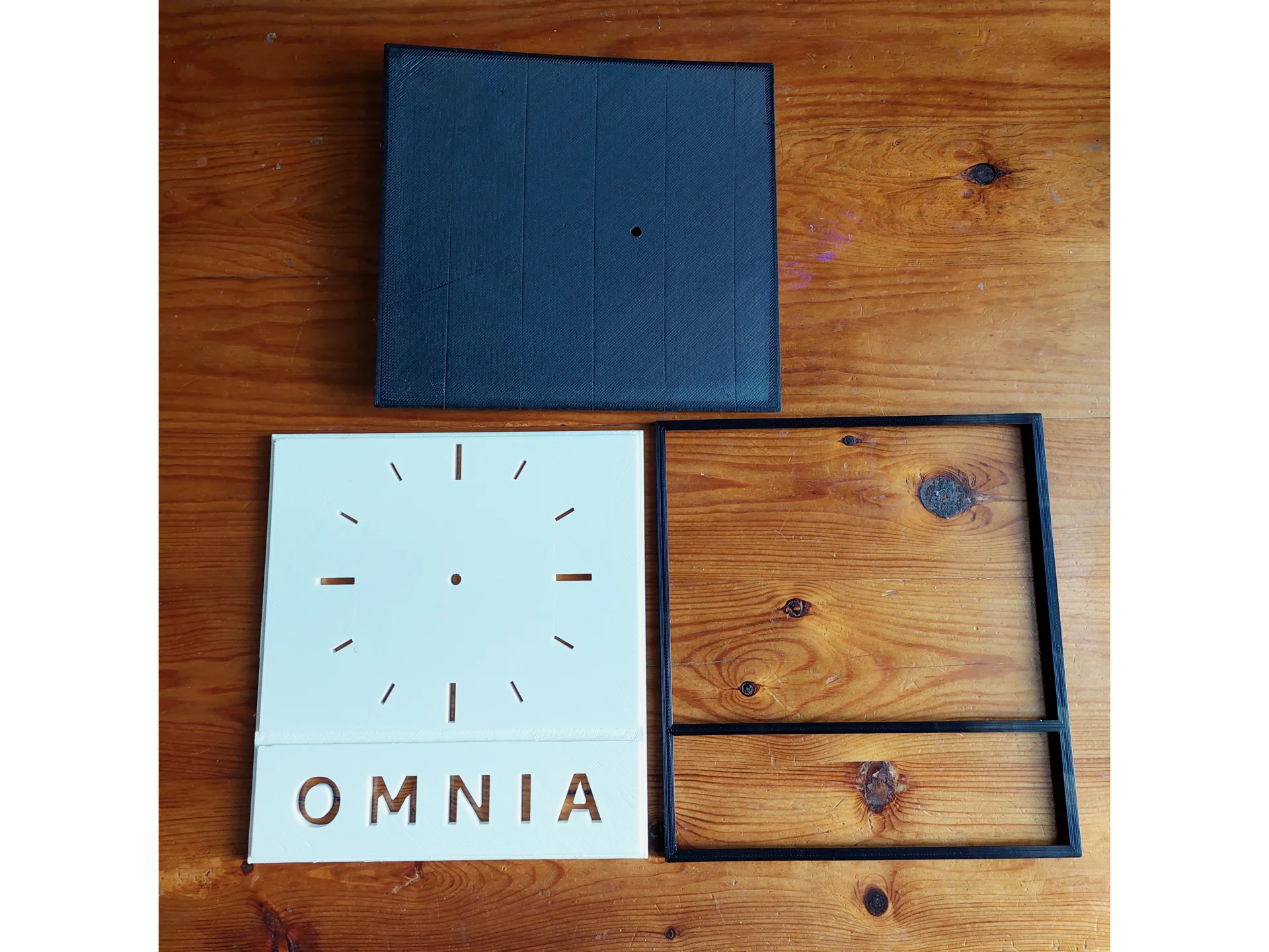 Omnia-logosta inspiroitu 3D-tulostettu seinäkello - osat, musta kehys, valkoinen taulu, musta tausta.