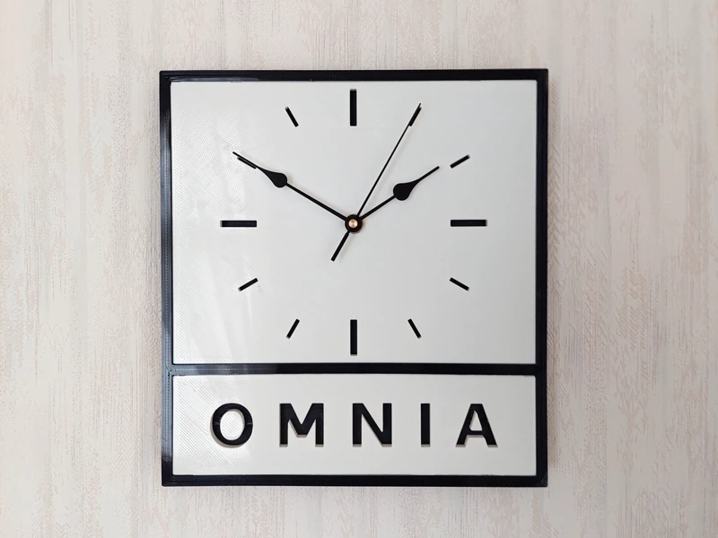 Omnia-logosta inspiroitu 3D-tulostettu seinäkello, musta kehys, valkoinen taulu, musta tausta.