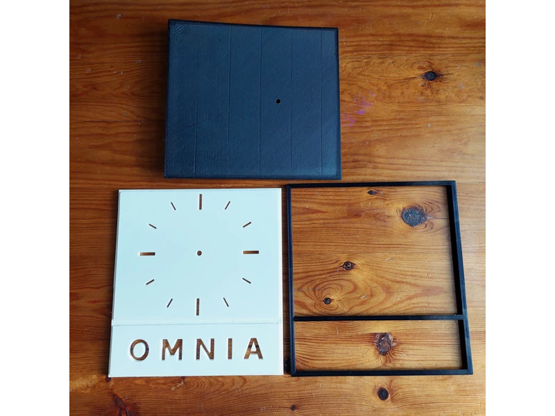 Omnia-logosta inspiroitu 3D-tulostettu seinäkello - osat, musta kehys, valkoinen taulu, musta tausta.